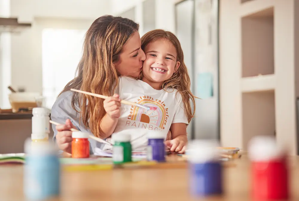 A mother kisses her smiling daughter as they paint together at a table with colorful paints nearby