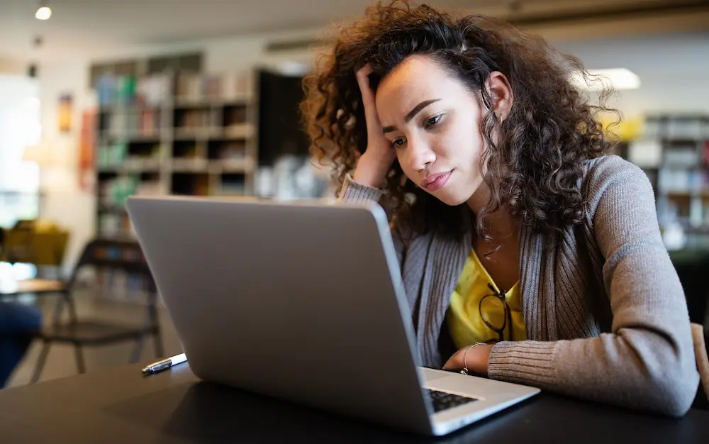 Stressed woman sitting at a laptop with her head resting on her hand appearing distracted and overwhelmed while working illustrating habitual procrastination